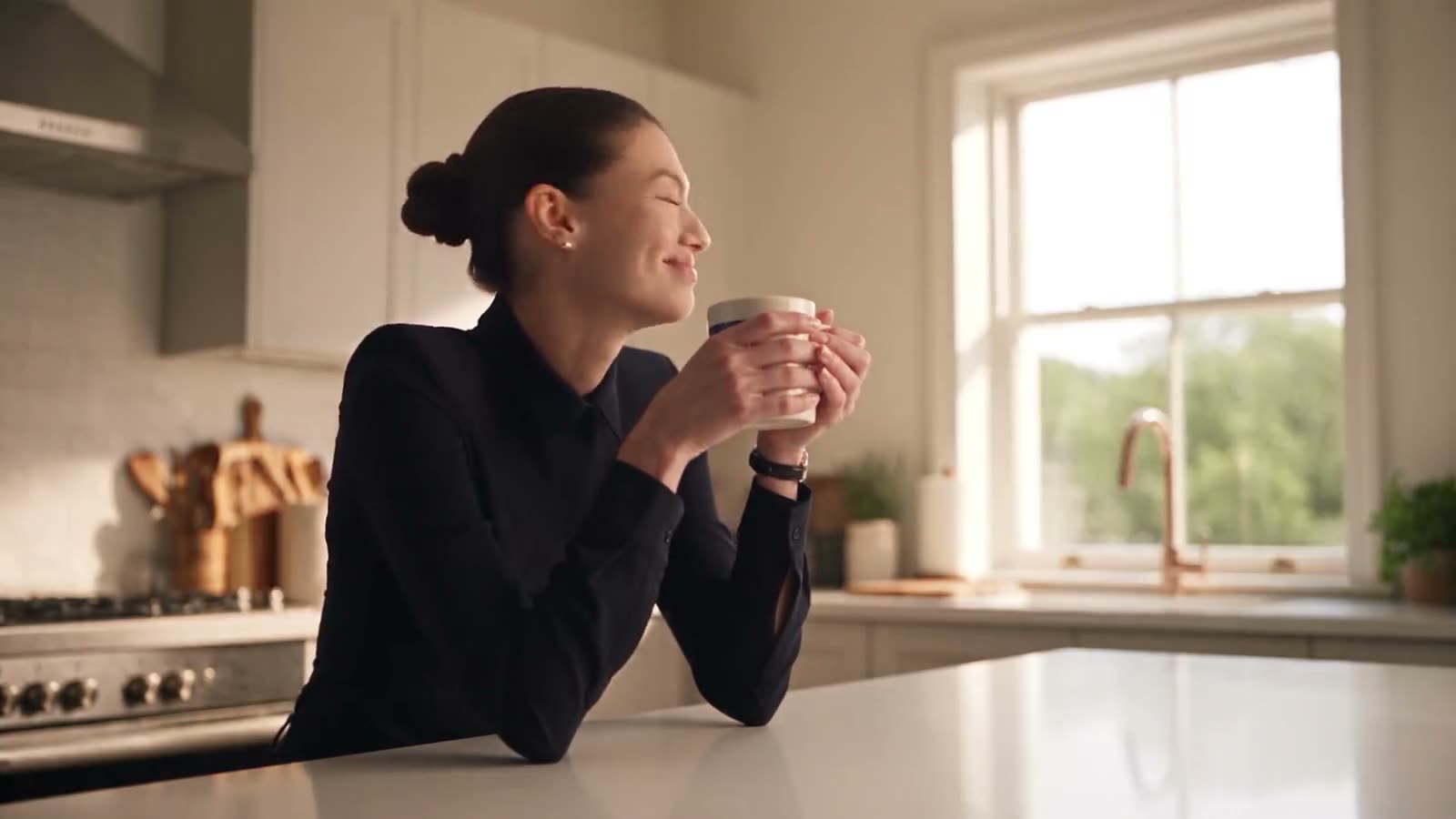 A woman leaning on a kitchen counter in morning light, eyes closed and smiling, holding a mug of coffee in both hands